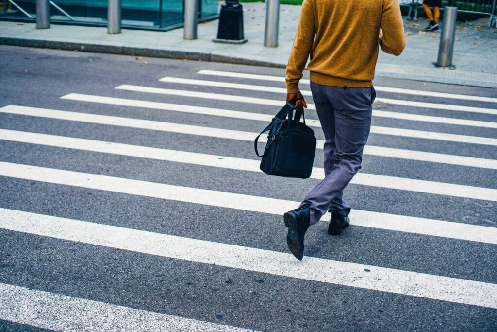 man walking across zebra crossing with a work bag in his hand wearing a yellow jumper and grey trousers