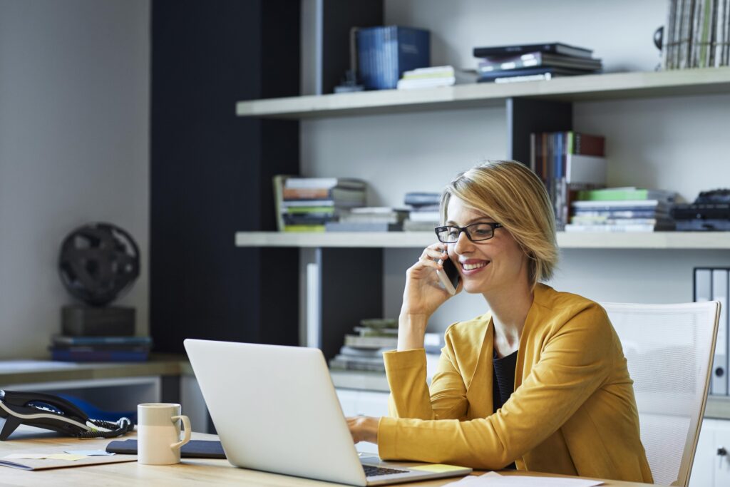 white woman wearing a yellow jacket with glasses and short blonde hair, sat at a desk on her laptop and mobile phone.